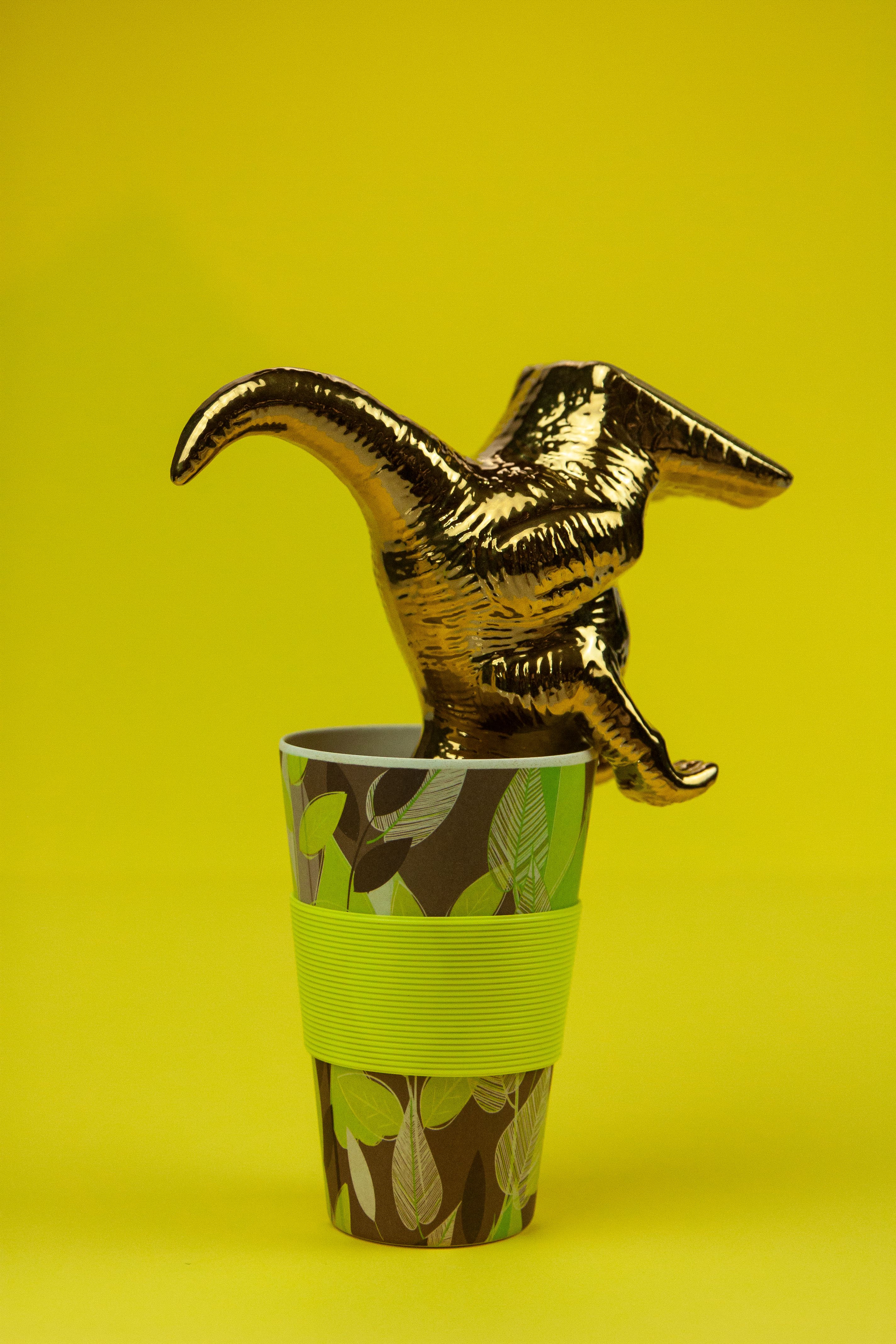 A golden dinosaur figurine displayed elegantly in a patterned paper cup against a vibrant yellow background.