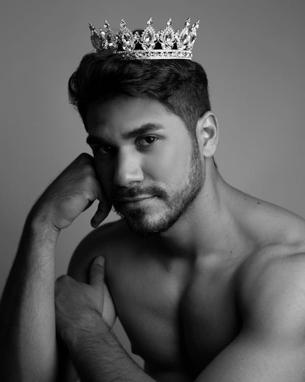 Black and white of muscular shirtless bearded ethnic male model with princess crown on head leaning on hand while sitting in studio and looking at camera