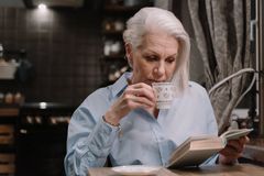 Elderly woman in a button-down shirt sipping tea while reading a book indoors.