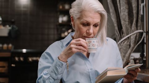 Elderly woman in a button-down shirt sipping tea while reading a book indoors.
