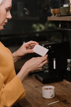 An elderly woman holds a note next to a cup of coffee and coffee machine indoors.