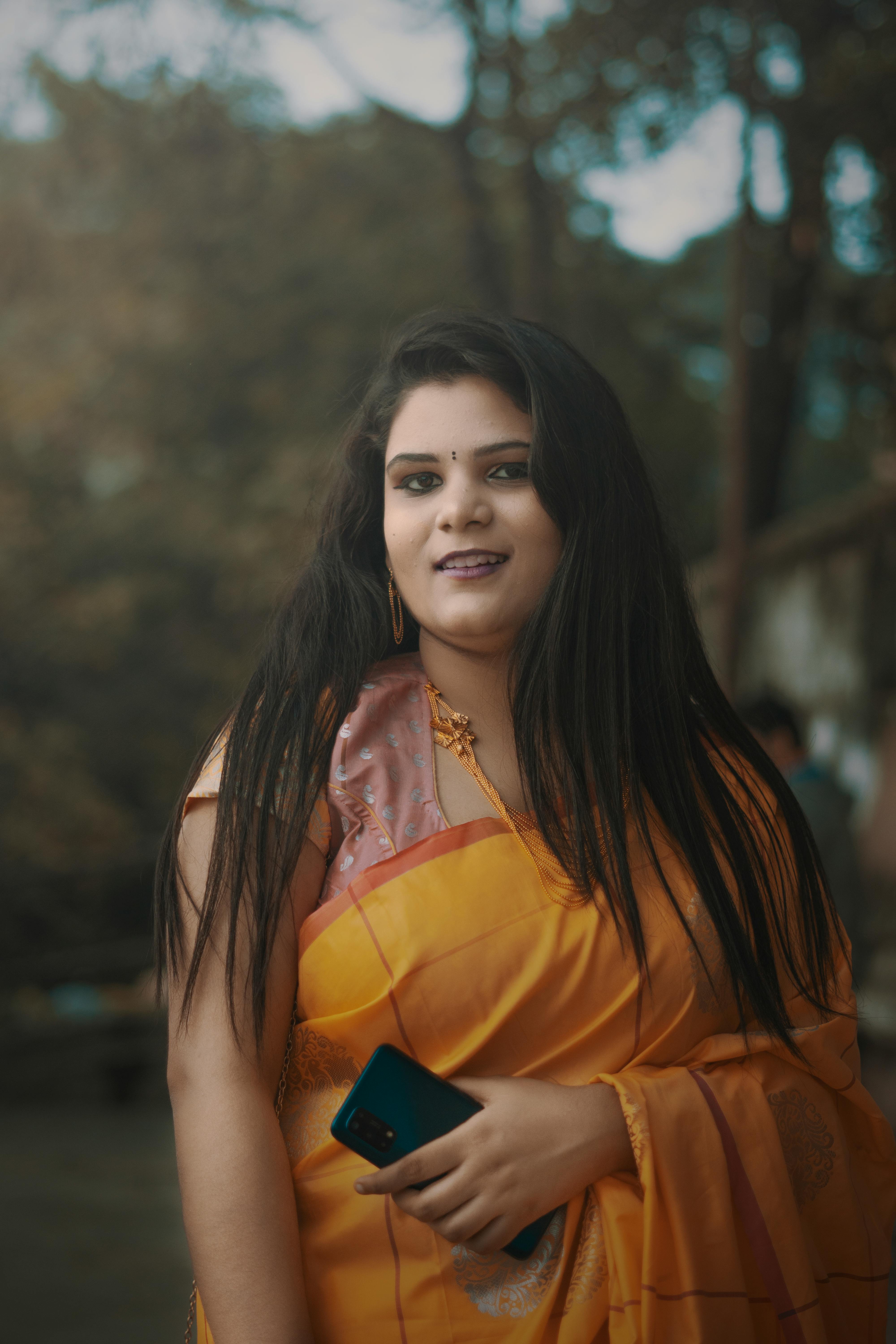 Young woman smiling outdoors, wearing a vibrant yellow sari, and holding a mobile phone.