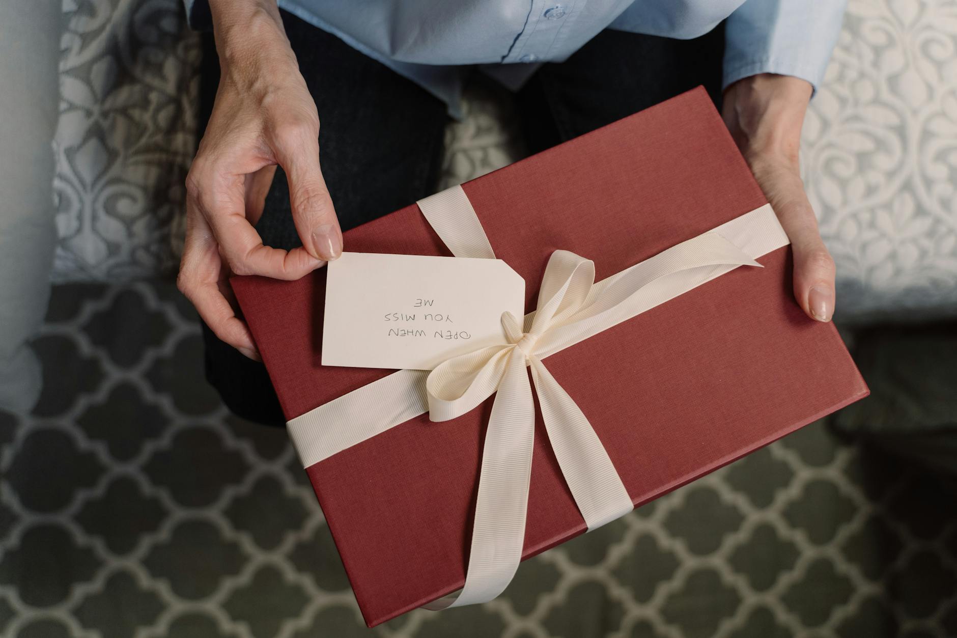 Close-up of hands holding a red gift box with a white ribbon and a message tag.
