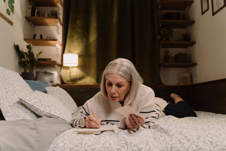 An Elderly Woman Lying On Bed Writing A Letter