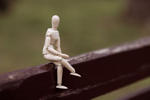 A wooden mannequin sits thoughtfully on a rustic fence, surrounded by nature's calm.