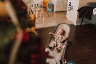 A cute baby in a bouncer seat gazes at a Christmas tree in a cozy living room setting.
