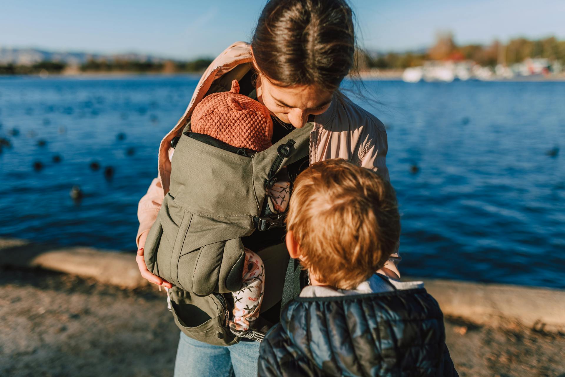 A mother holding her baby in a carrier while her young son stands nearby by a scenic waterfront.