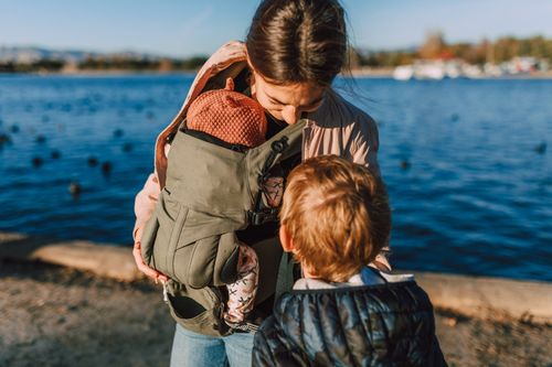 Free A mother holding her baby in a carrier while her young son stands nearby by a scenic waterfront. Stock Photo