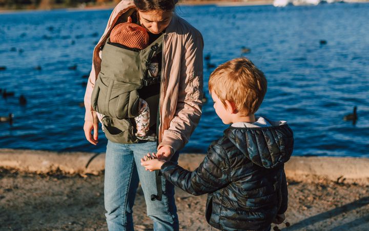 A mother with her child and baby carrier walks by a serene lakeside on a sunny day.