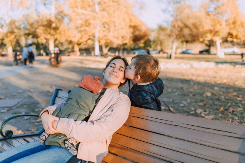 A smiling mother sitting on a park bench with her toddler and baby