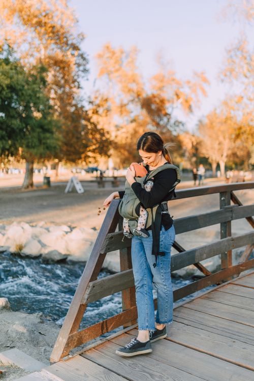 Free Woman holding baby in a carrier while standing on a wooden bridge in a park during autumn. Stock Photo