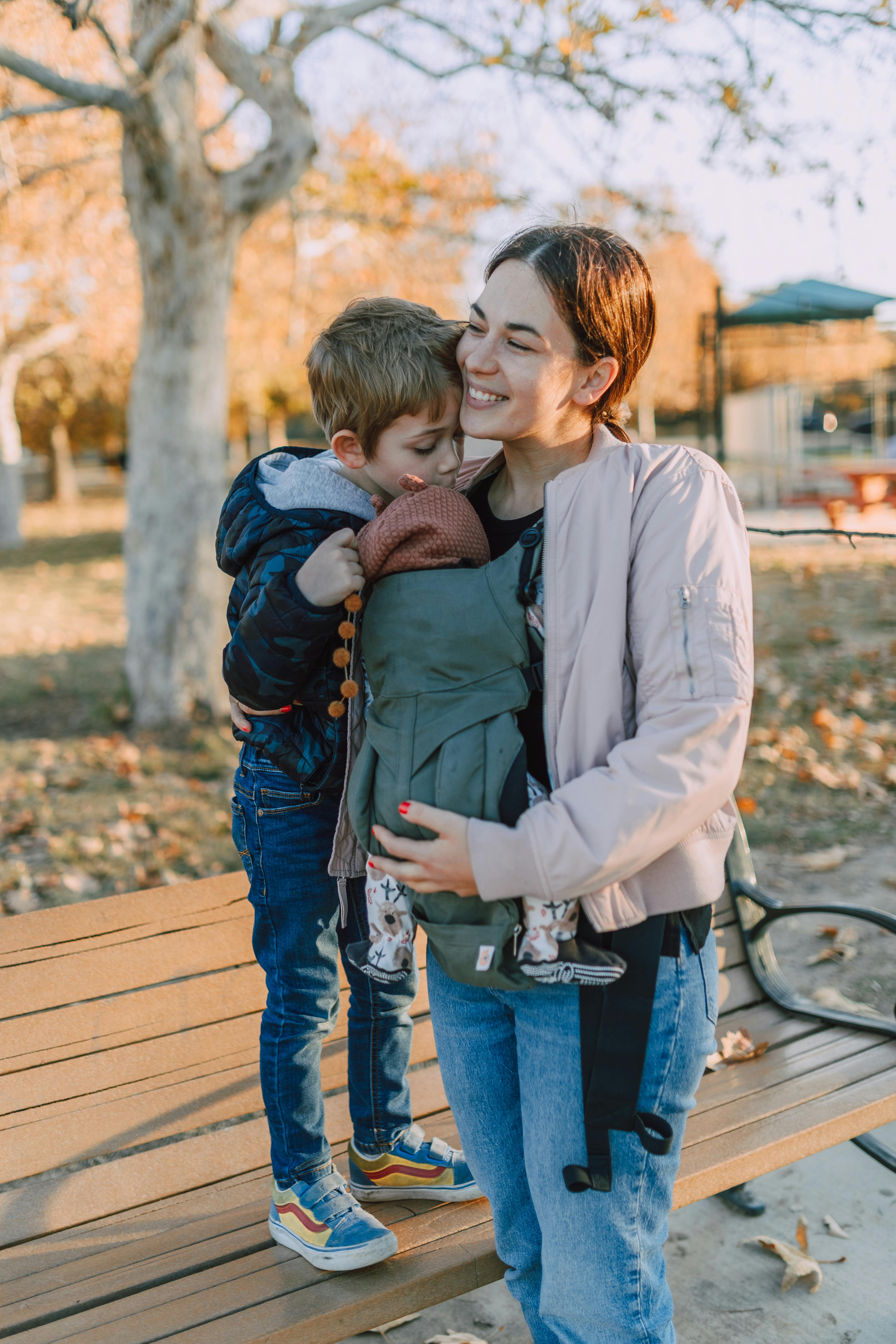 a young boy standing on a wooden bench while looking at the baby carried by his mother