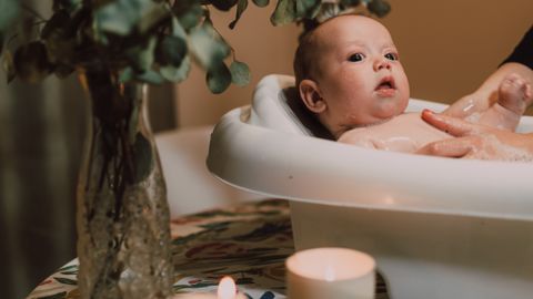 A cute baby in a tub surrounded by candles and greenery on a table indoors.