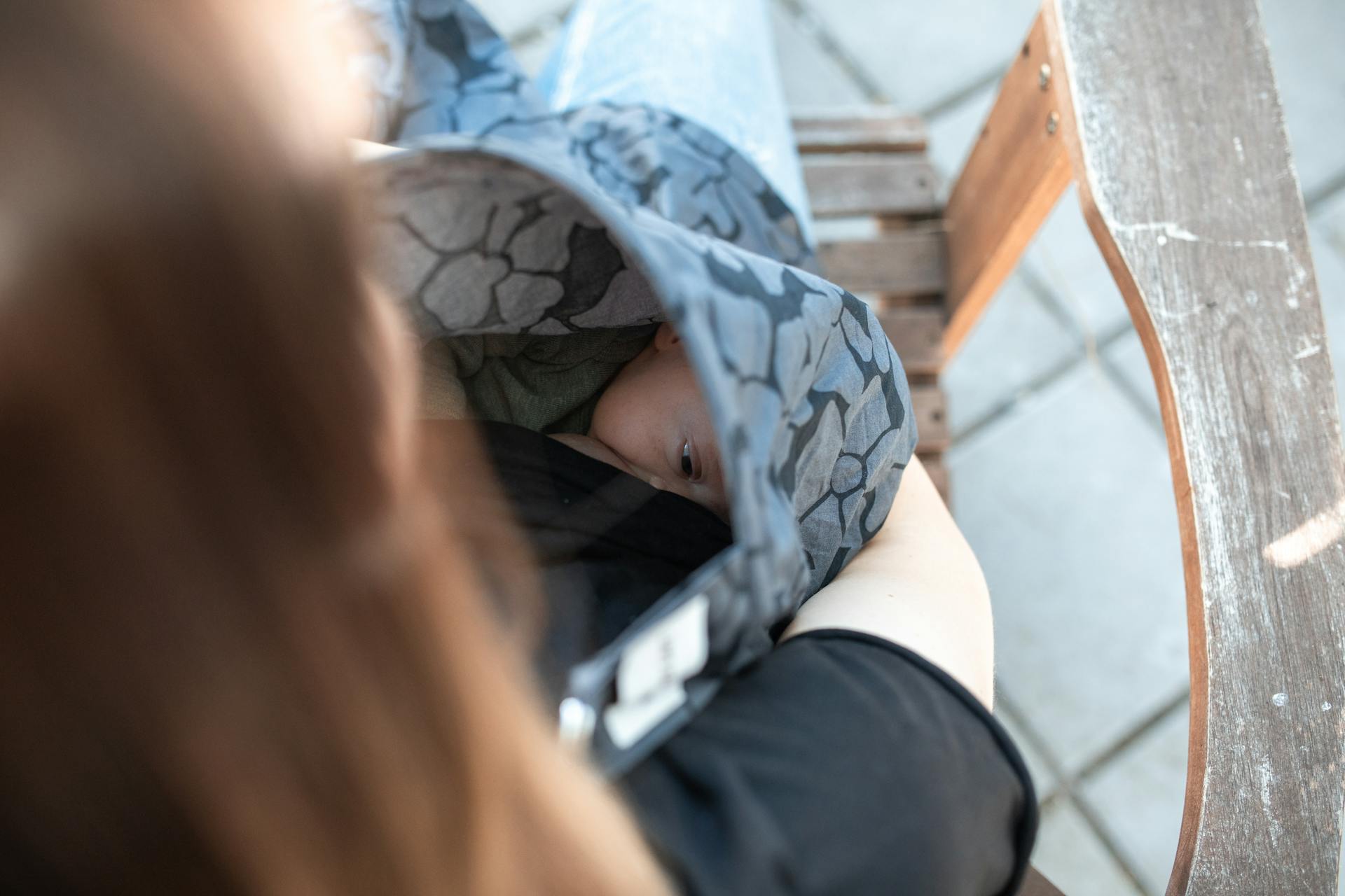 Intimate outdoor moment of mother breastfeeding baby under cover on a bench.