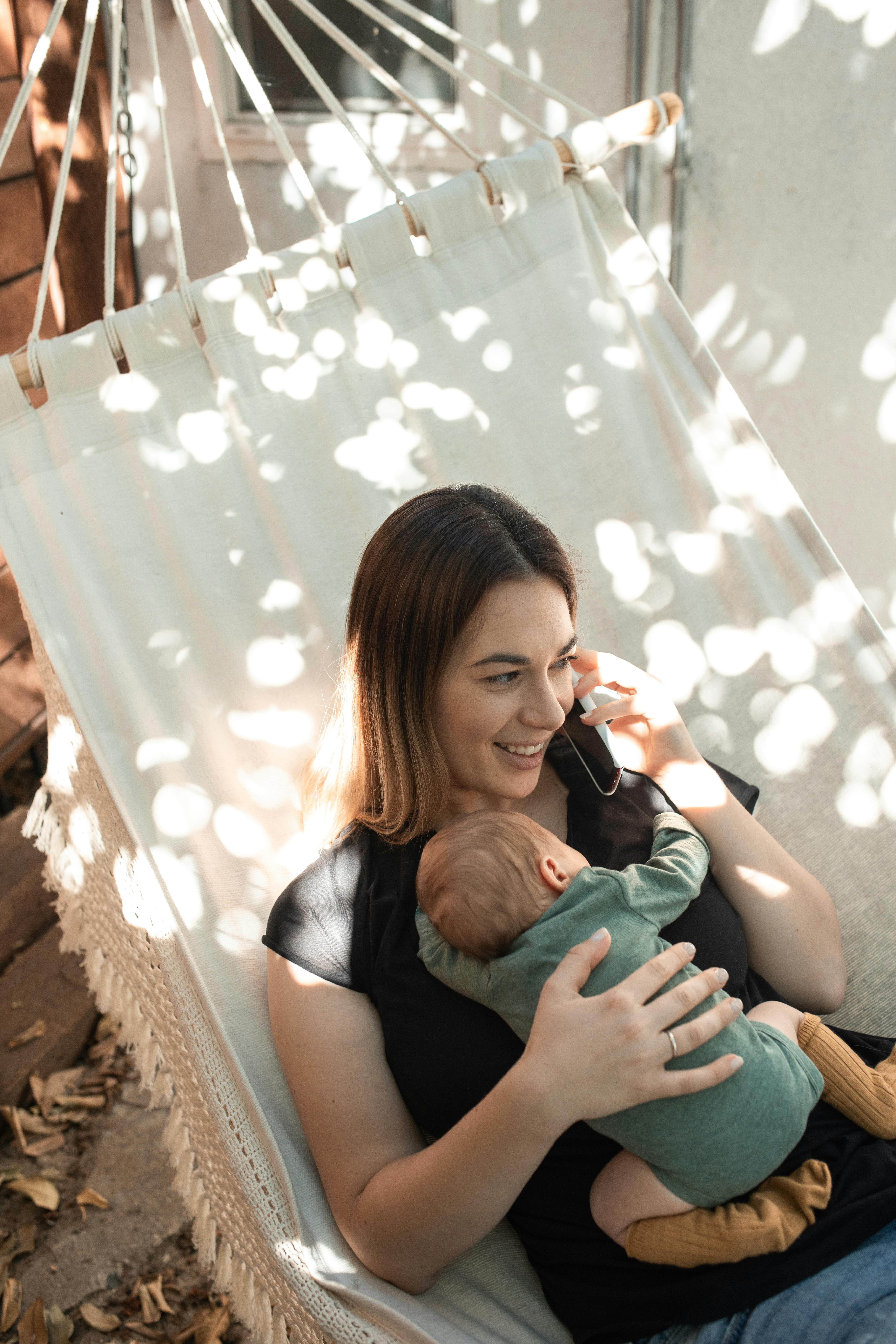 A mother relaxes in a hammock with her newborn, chatting on the phone in a serene indoor setting.
