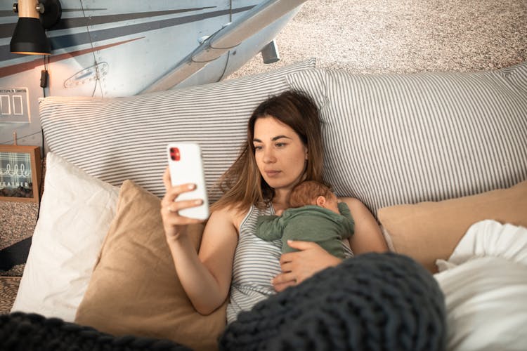 A Woman Lying Down With Her Baby On Her Chest While Using Her Mobile Phone