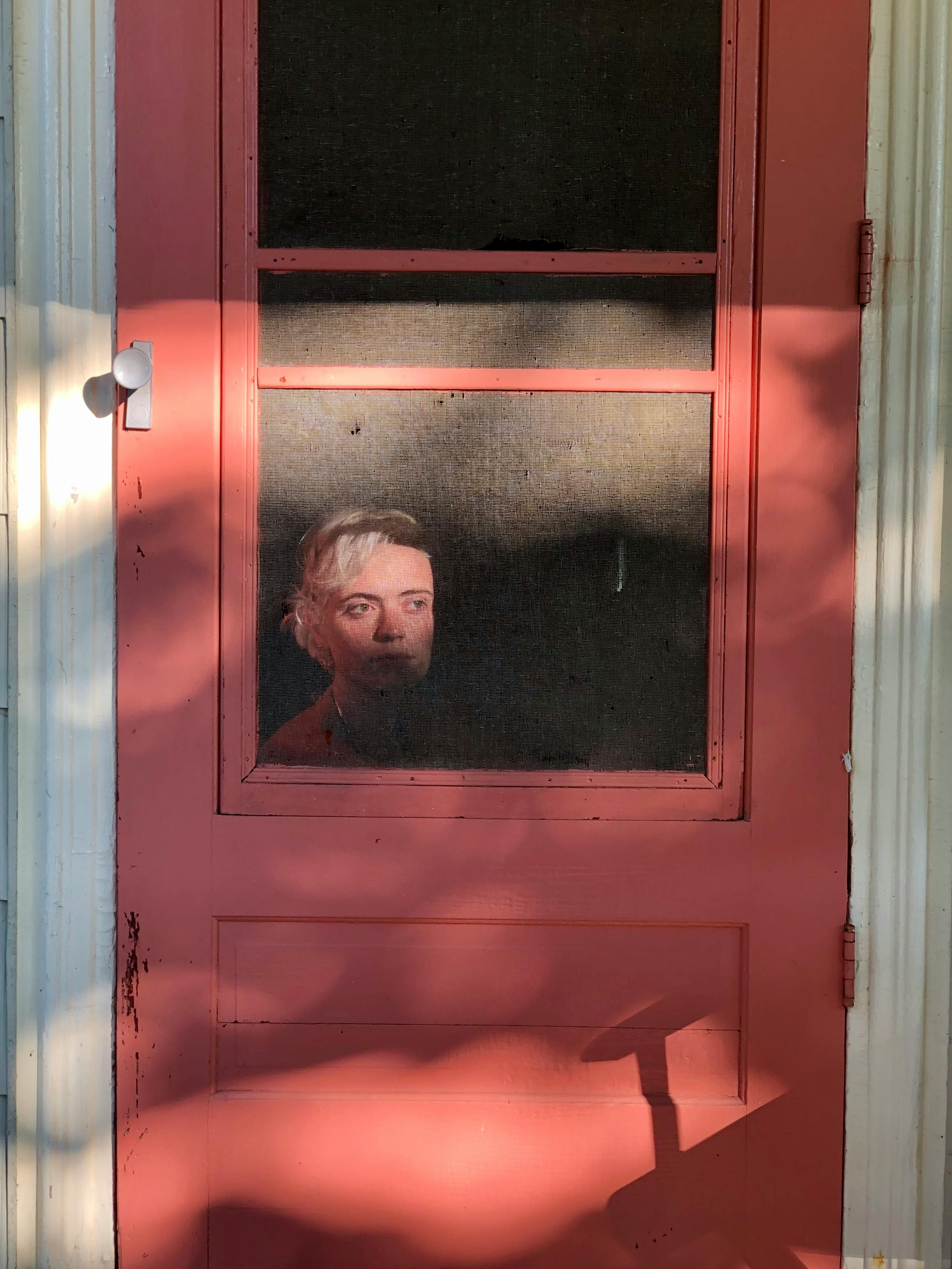 Contemplative young female sitting behind red door of rural house and looking away in thoughts
