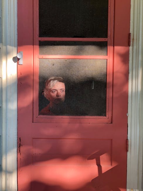 Contemplative young female sitting behind red door of rural house and looking away in thoughts