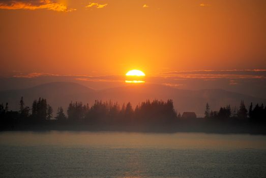 Scenic view of bright orange sun setting over hilly terrain located near calm rippling river