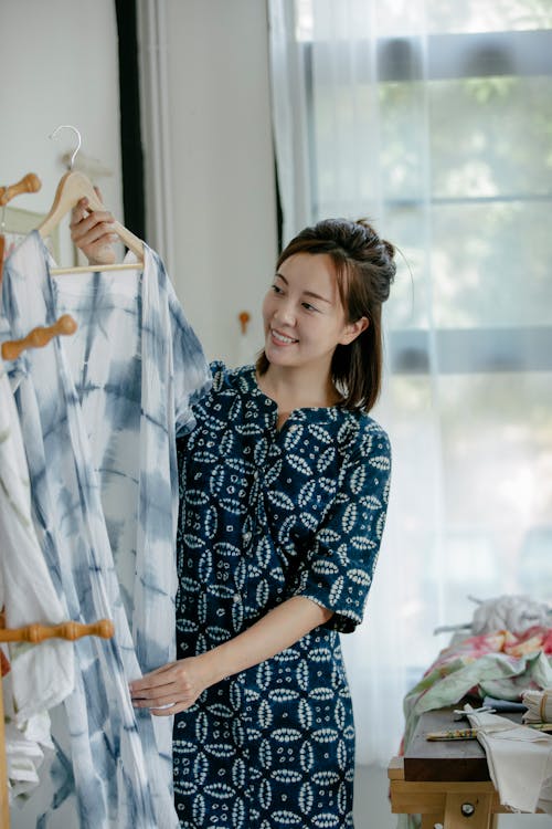 Positive Asian female dressmaker with long clothes on hanger standing near rack while working in sewing workshop