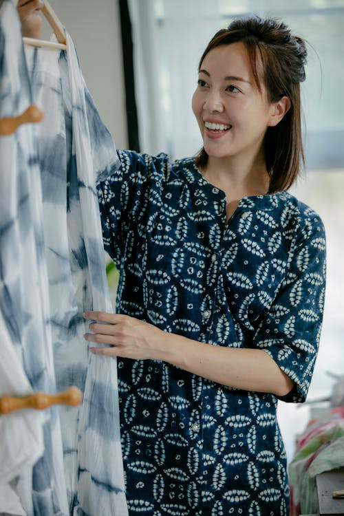 Positive Asian female designer with toothy smile taking hanger with clothes in sewing workshop