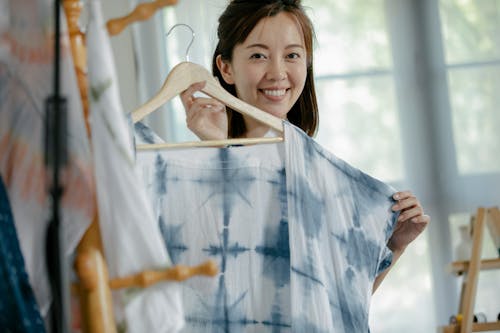Positive professional Asian female designer with modern garment on hanger looking at camera while standing near clothes in modern studio