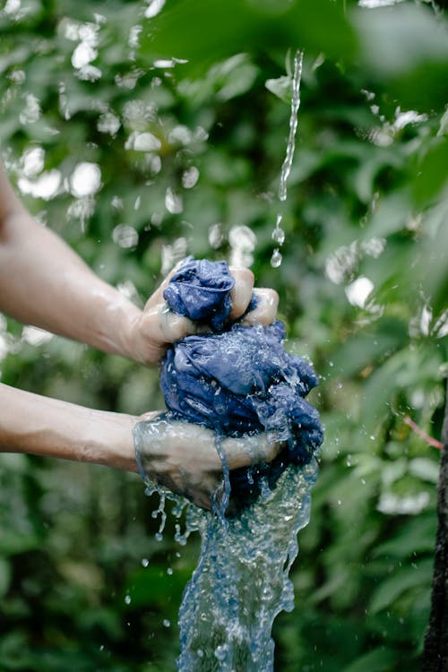 Hands of unrecognizable person squeezing wet blue dyed textile under water while standing on street with green plants on blurred background