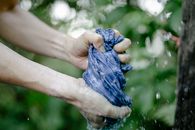 Hands of faceless person squeezing wet blue fabric under water drops while standing on street near green plants on blurred background