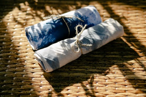 From above rolls of dyed white and blue textile tied with ropes placed on wicker surface in professional workshop with sunlight