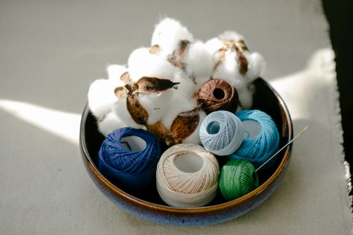 Free From above of sunlit clay bowl with colorful yarn for handicraft and white balls of cotton fiber placed on beige cloth on table Stock Photo
