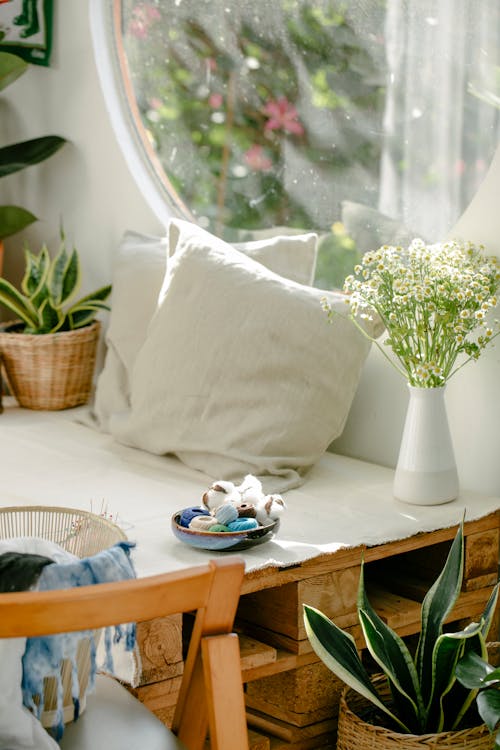 Free Vase of fresh gentle chamomiles and potted plant placed on table near cushions and bowl with various thread spools in cozy atelier Stock Photo
