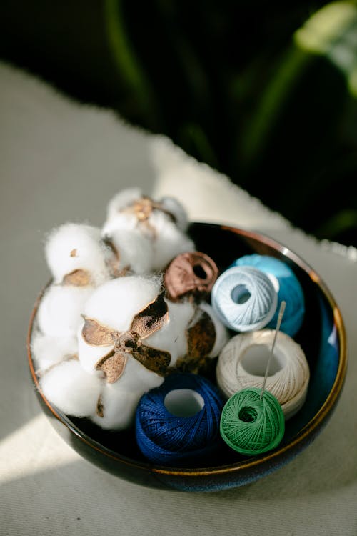 From above of set of colorful silk thread bobbins placed in bowl with natural cotton balls in atelier