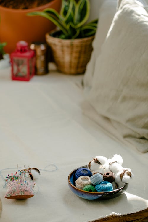 From above of bowl with colorful thread bobbins and cotton balls placed on table near small pillow with pins and needles in light atelier