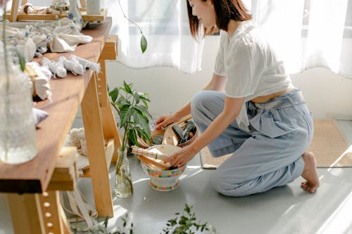 Side view of crop happy young ethnic female designer sitting on haunches and choosing tools for tie dyeing clothes with shibori technique in workshop