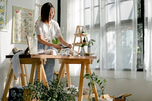 Positive young Asian female designer cutting clothes while standing at wooden table in creative workshop decorated with various potted plants