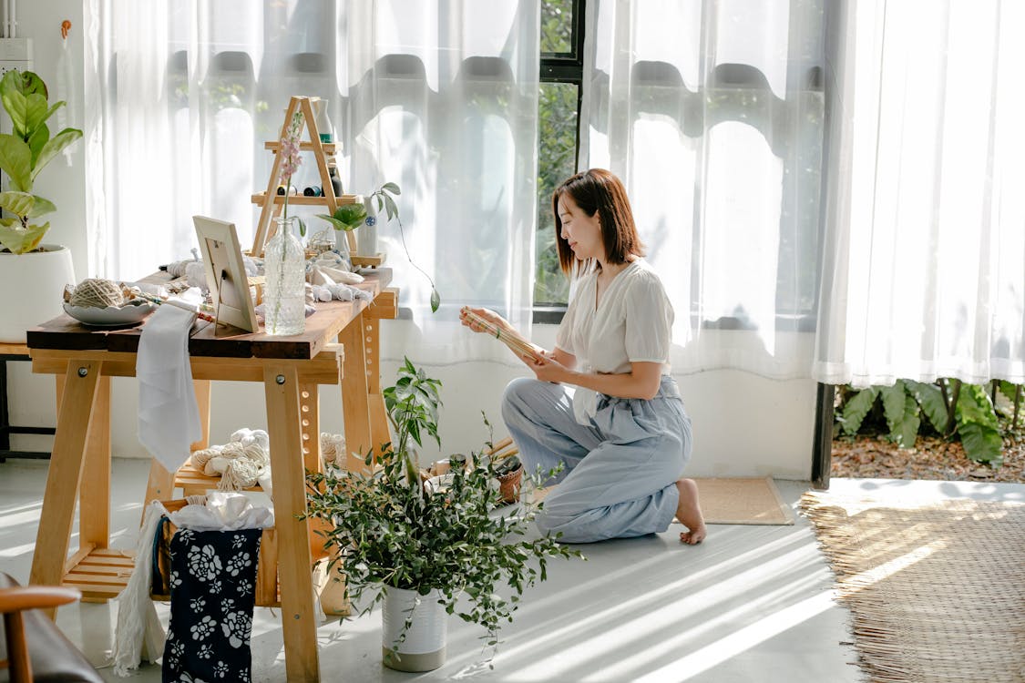 Free Side view of happy young barefooted female tailor siting on haunches in atelier near wooden table with heap of various fabrics in sunlight Stock Photo