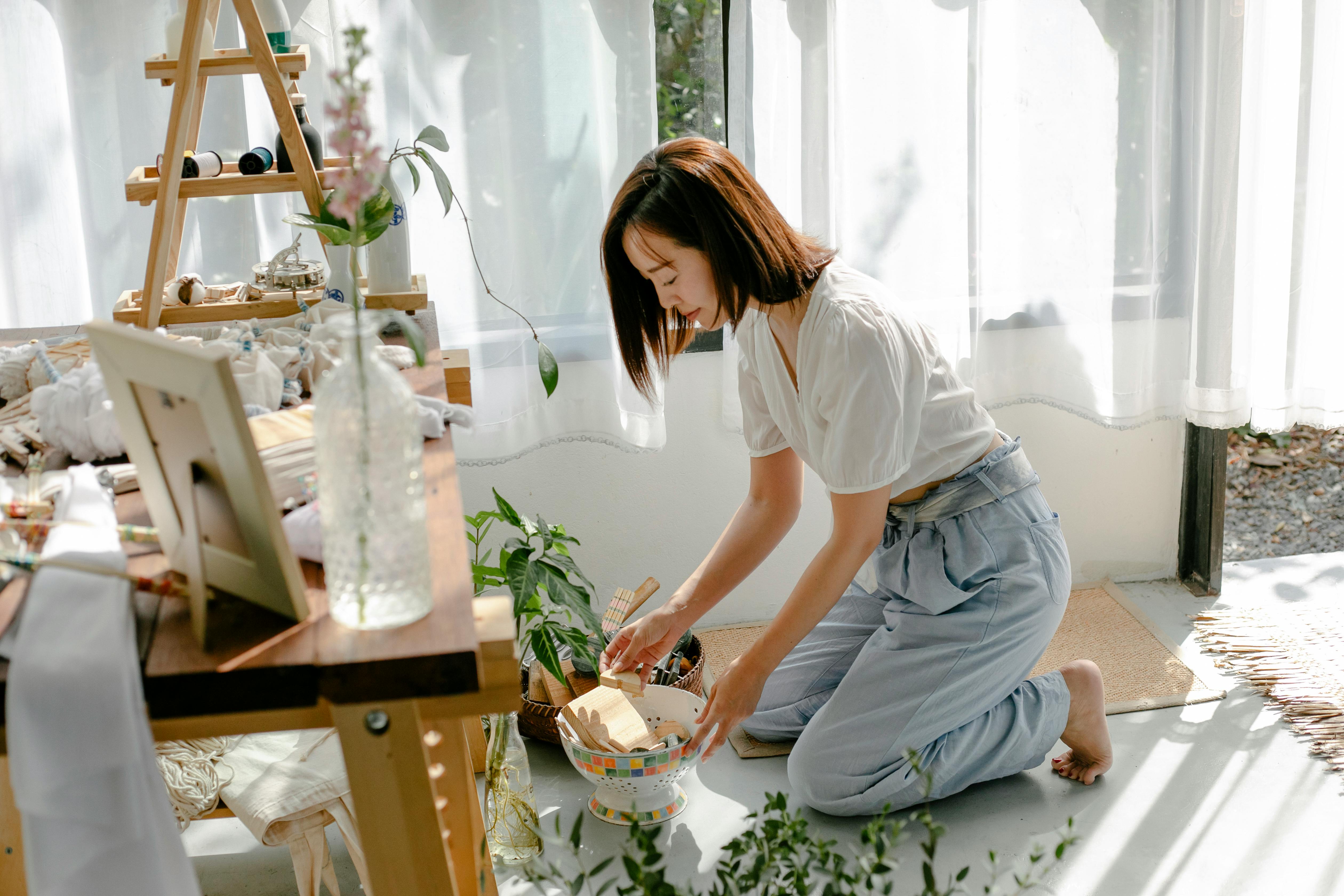 Side view full body of barefooted young ethnic female sitting on knees on floor while choosing tools for dyeing clothes with shibori technique