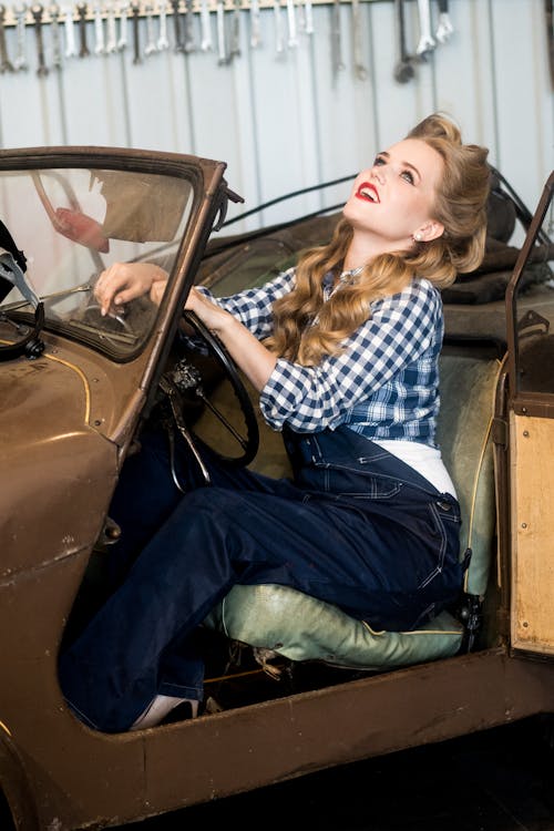 Confident young blond haired woman in overall and shirt sitting in vintage car with open door in light garage near instruments