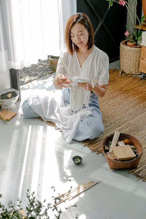 Asian craftswoman wrapping cotton fabric around poles while showing traditional Japanese tie dye technique in house on sunny day