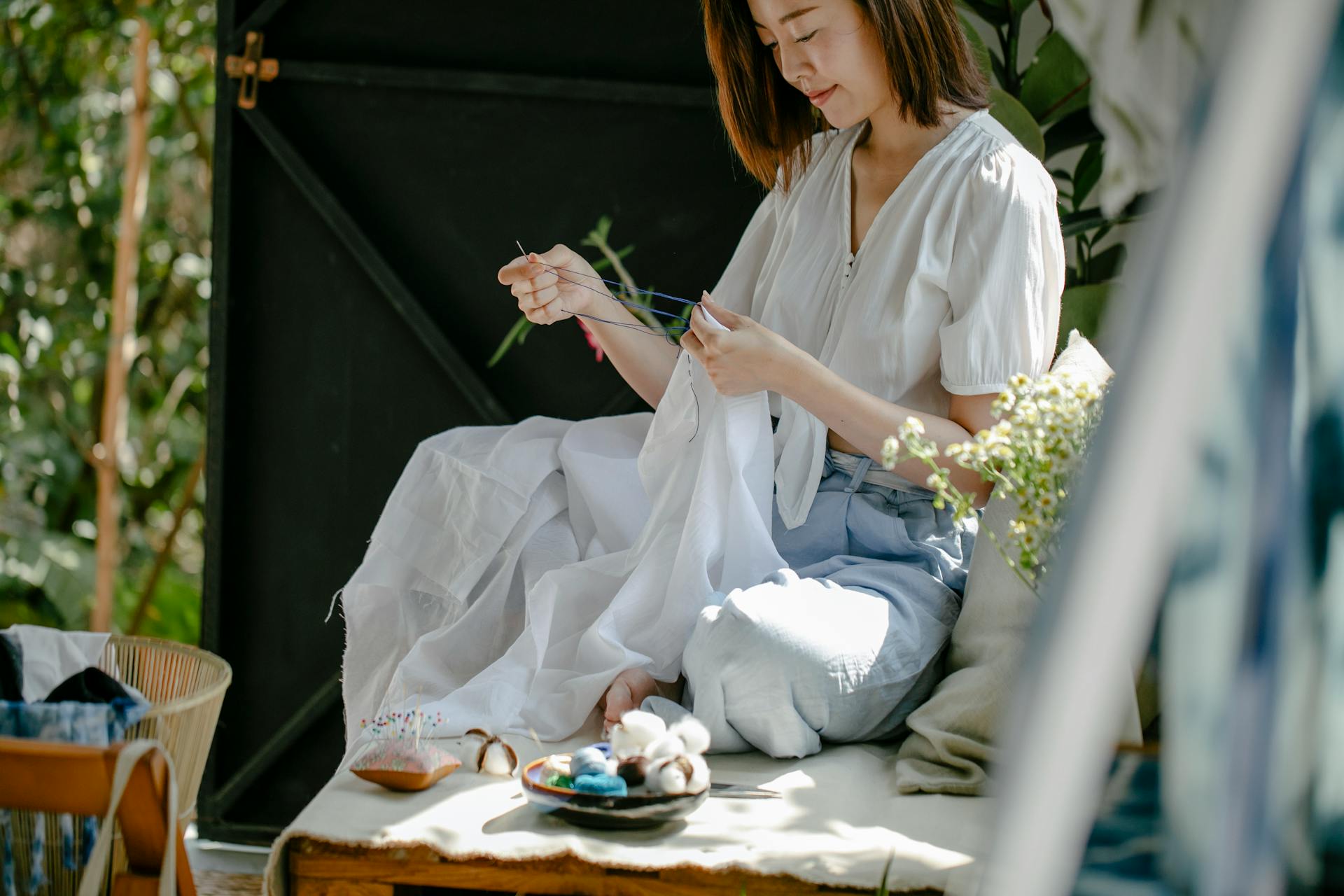 Crop ethnic craftswoman stitching textile with needle and thread while demonstrating traditional Japanese tie dye technique in sunshine