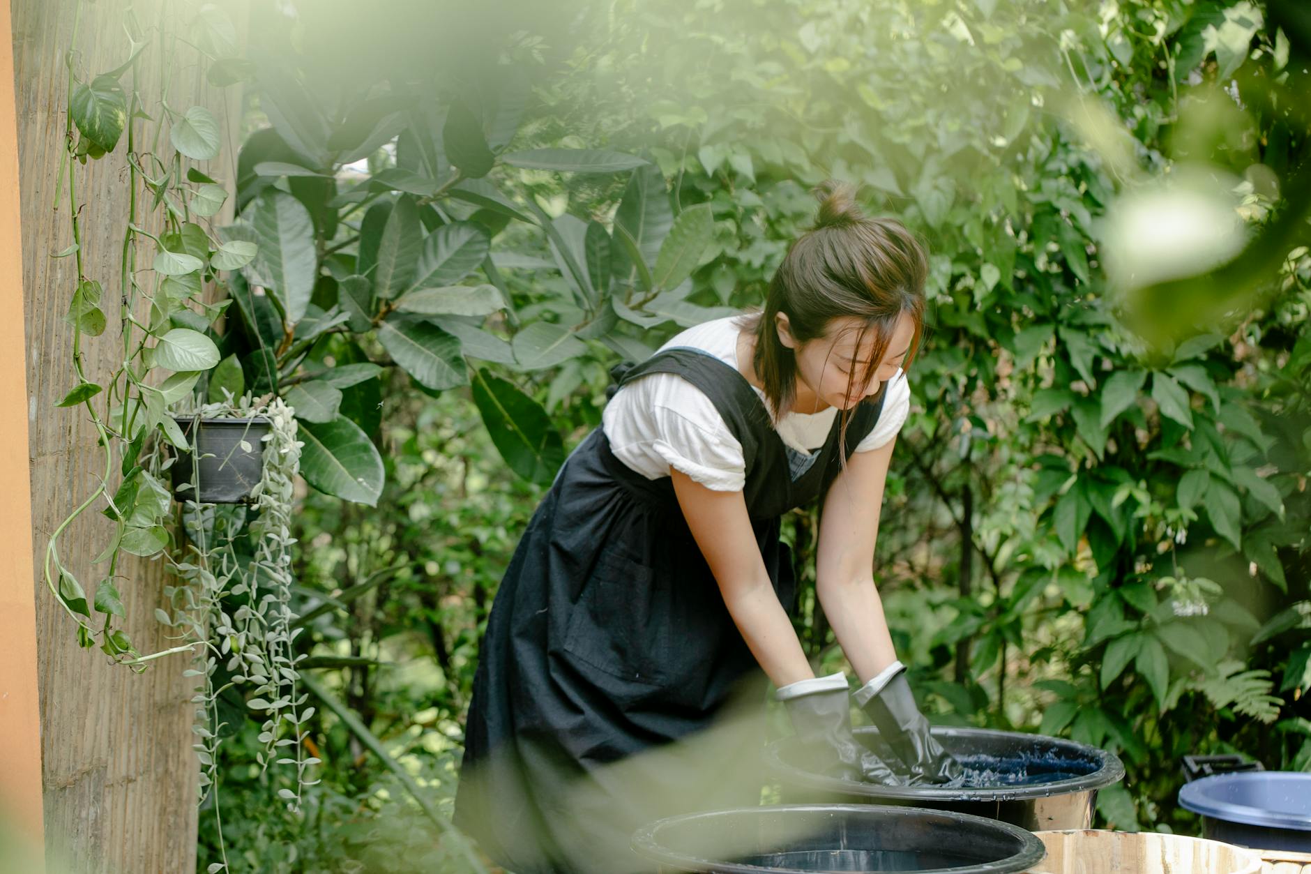 Young ethnic female artisan in gloves demonstrating shibori technique while painting cloth in a basin with water in a garden.