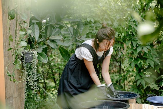 Young ethnic female artisan in gloves demonstrating shibori technique while painting cloth in basin with water in garden