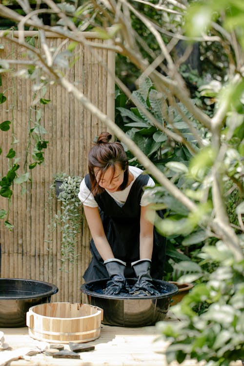 Asian craftswoman in gloves painting textile in basin with aqua while showing shibori technique in garden