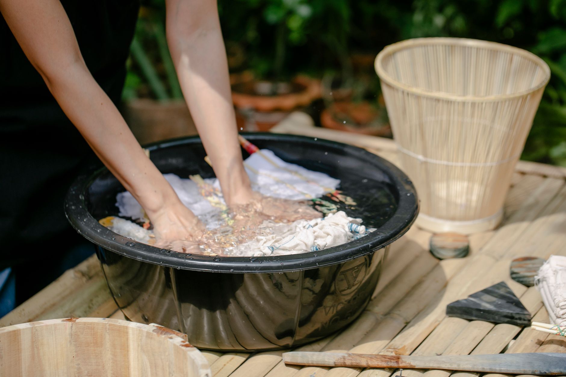 Crop unrecognizable artisan immersing textiles with poles in pure aqua in basin while demonstrating tie dye technique outdoors