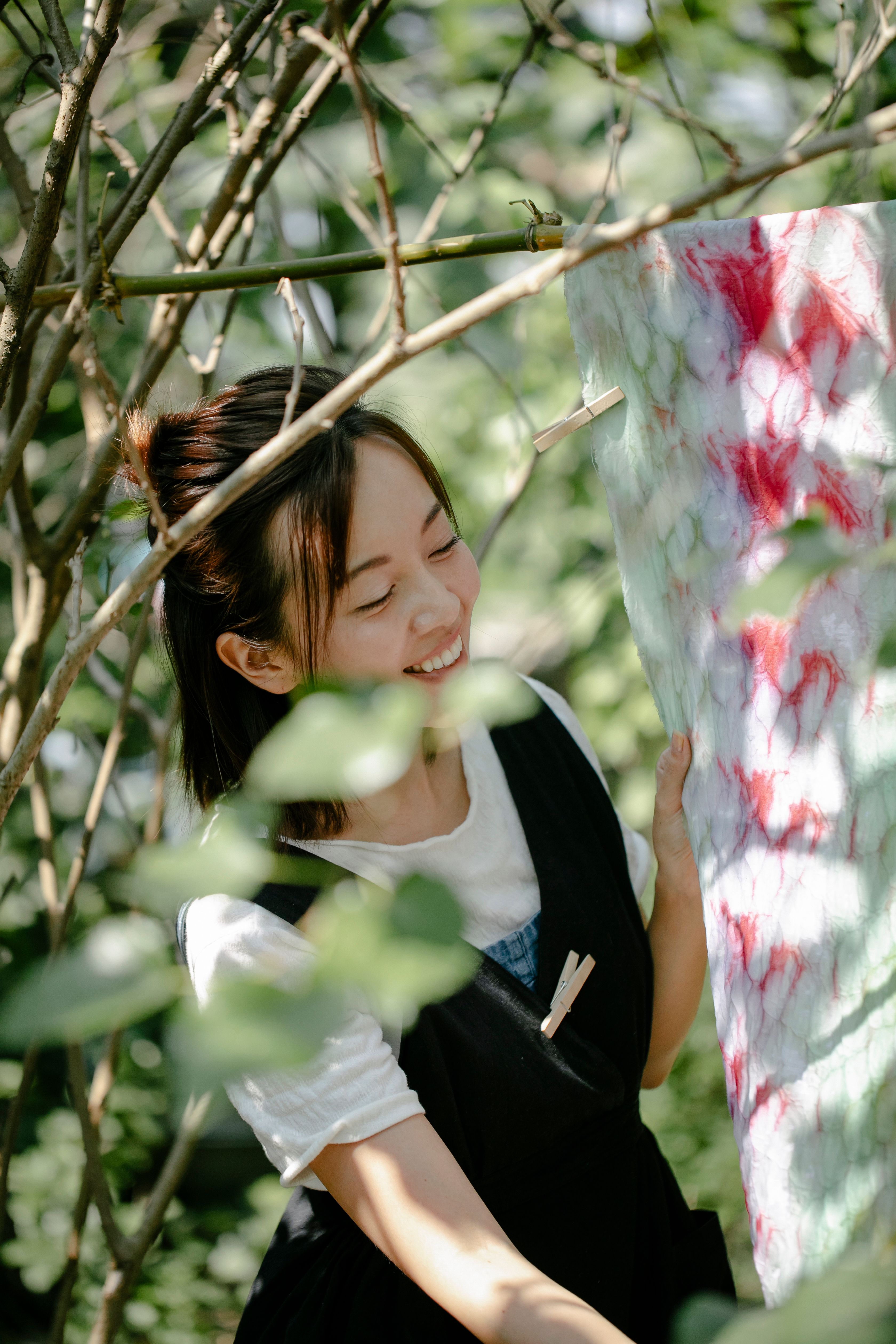 A woman smiling while hanging a colorful tie-dye cloth on a tree branch outdoors.