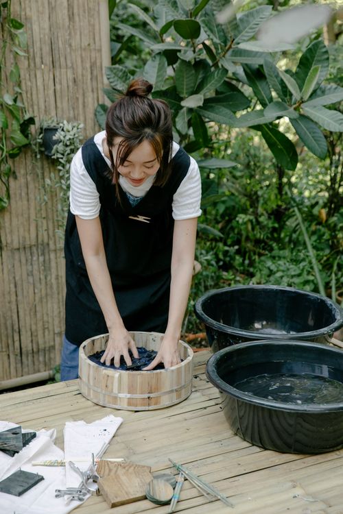 Free Asian woman standing outdoors tie-dyeing fabric, surrounded by greenery and natural elements. Stock Photo