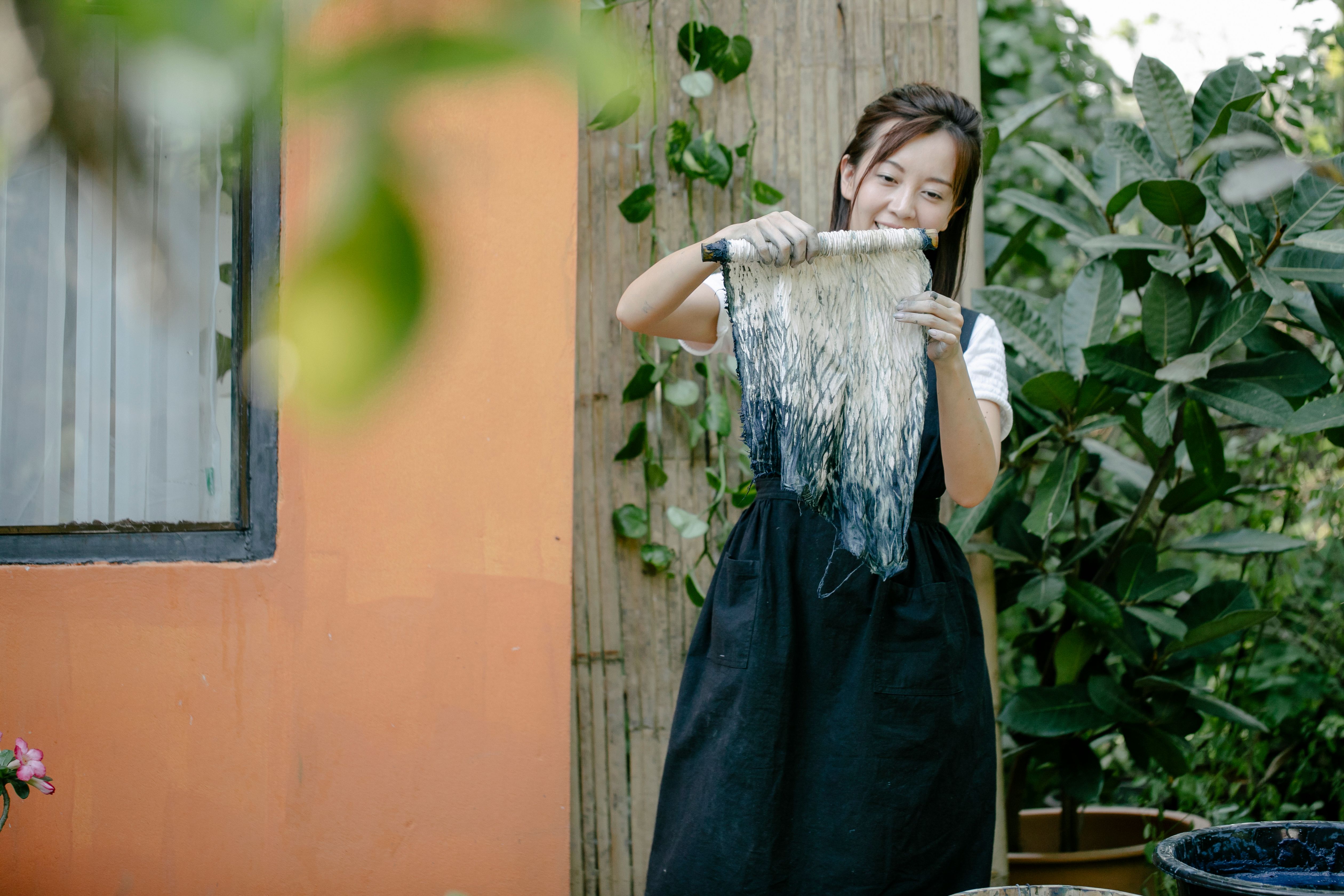 A woman smiling while holding a tie-dyed fabric outdoors in a garden setting, showcasing her creative work.