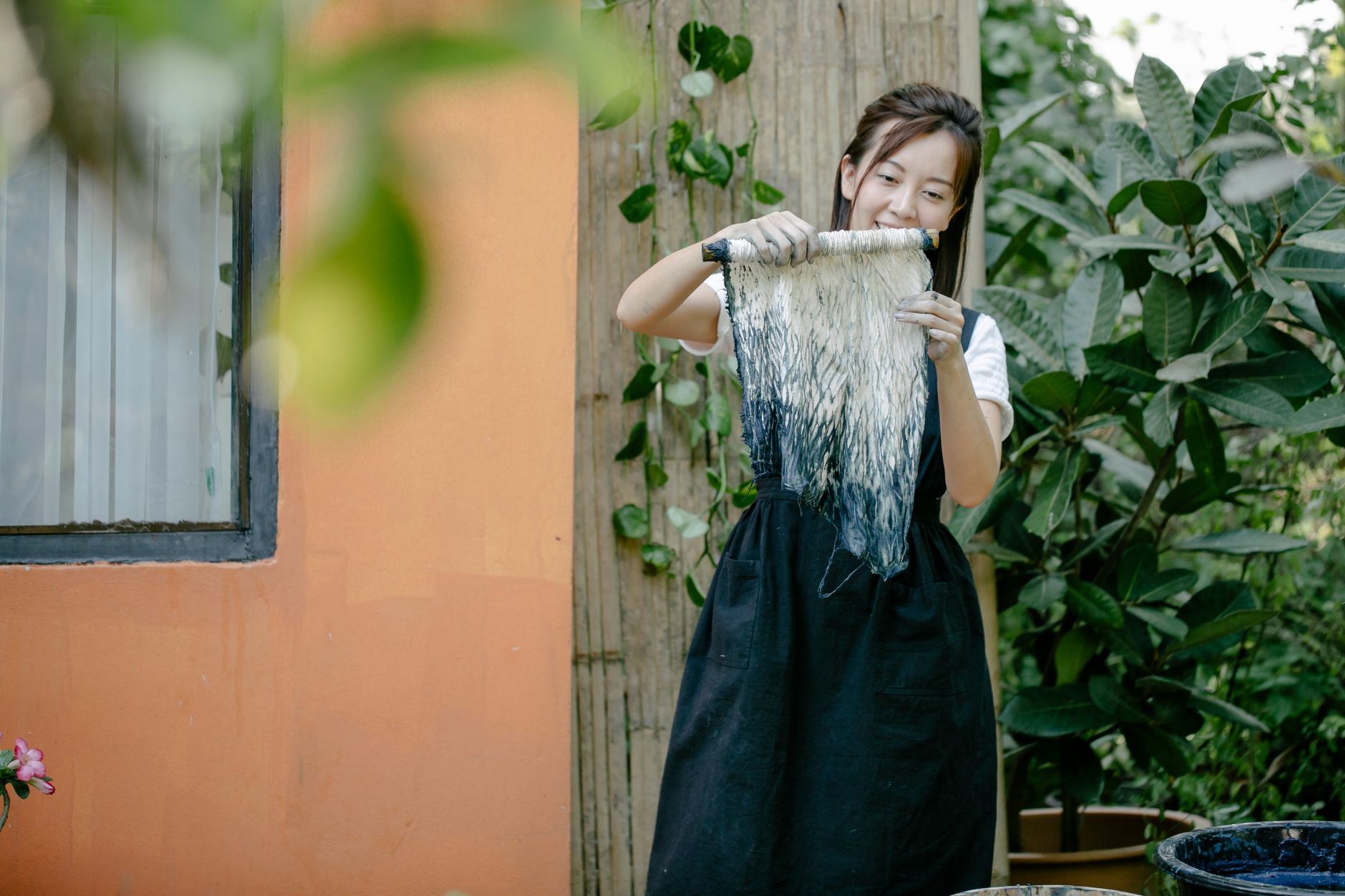 A woman smiling while holding a tie-dyed fabric outdoors in a garden setting, showcasing her creative work.