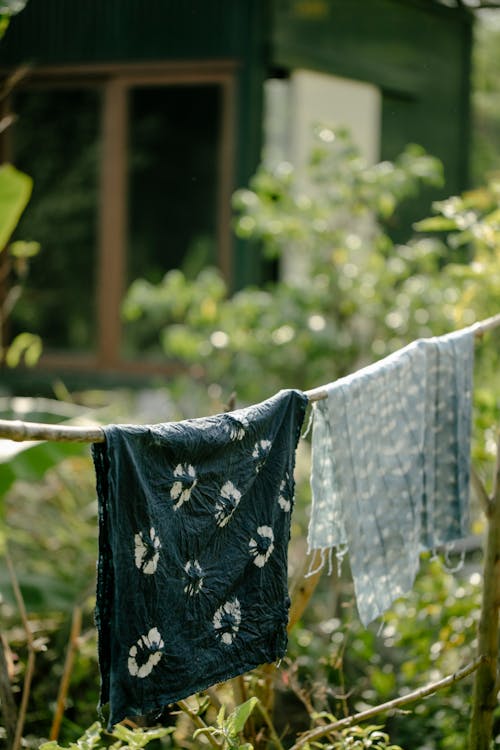 Painted textile with creative patterns using shibori technique hanging on rope in countryside with green plants against rural house on blurred background