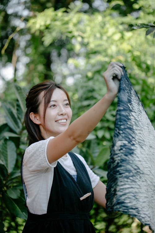 Happy Asian female in uniform with bright dyed textile looking away while standing near green plants on blurred background in countryside